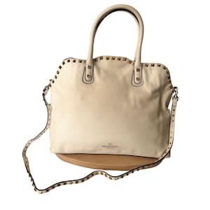 Puede incluir: Bolso de mano de cuero color crema con tachuelas doradas. Cuenta con un asa superior y una correa de hombro desmontable, ambas adornadas con tachuelas. El bolso tiene forma rectangular y el logotipo de Valentino.