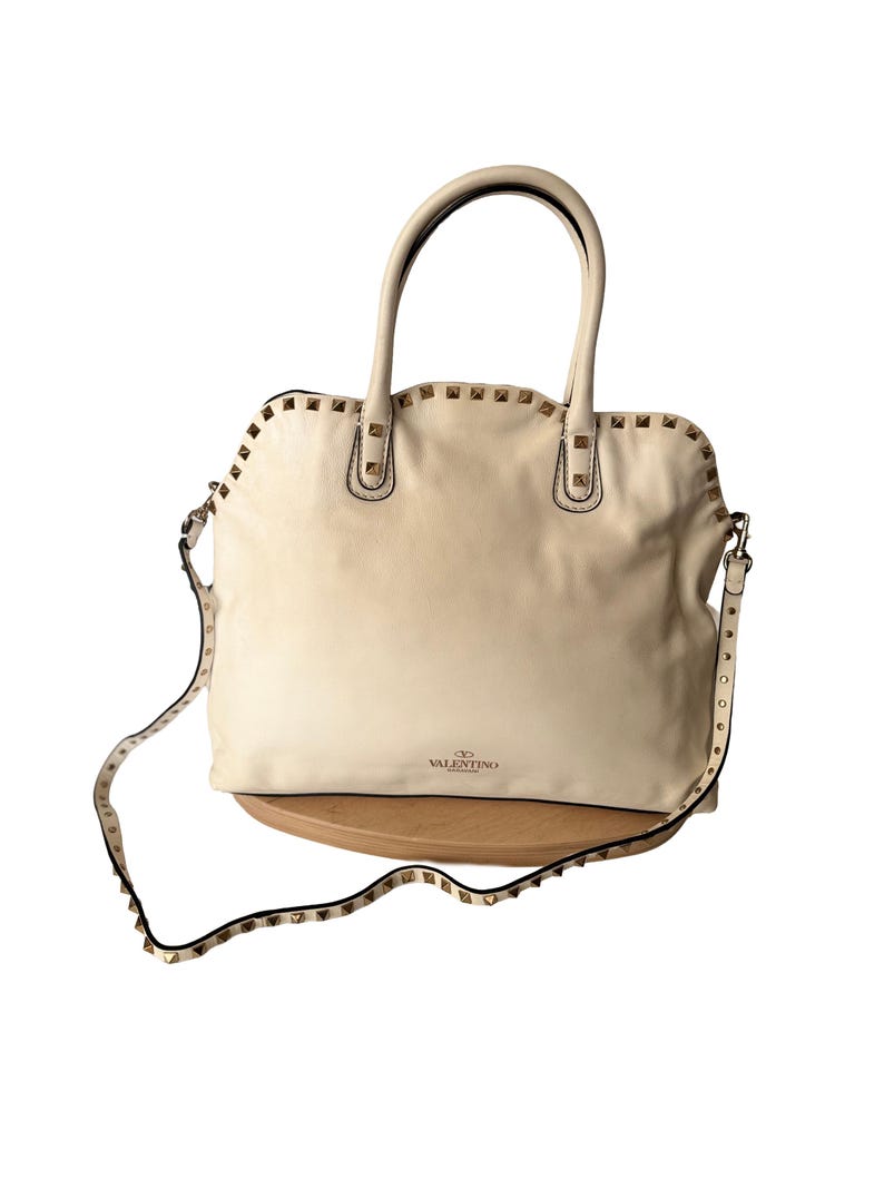 Puede incluir: Bolso de mano de cuero color crema con tachuelas doradas. Cuenta con un asa superior y una correa de hombro desmontable, ambas adornadas con tachuelas. El bolso tiene forma rectangular y el logotipo de Valentino.