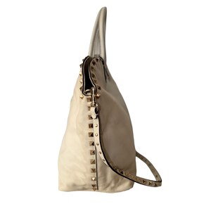 Puede incluir: Bolso de mano de cuero color crema con tachuelas doradas. El bolso tiene una correa para el hombro y un dise&ntilde;o holgado. Las tachuelas est&aacute;n dispuestas a lo largo de los lados y la correa, agregando un elemento decorativo al bolso.