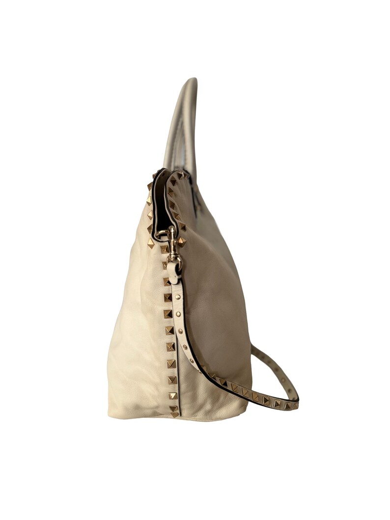 Puede incluir: Bolso de mano de cuero color crema con tachuelas doradas. El bolso tiene una correa para el hombro y un dise&ntilde;o holgado. Las tachuelas est&aacute;n dispuestas a lo largo de los lados y la correa, agregando un elemento decorativo al bolso.