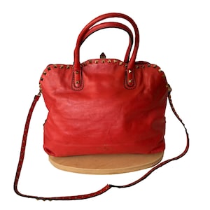 Peut inclure: Sac &agrave; main en cuir rouge vif orn&eacute; de clous dor&eacute;s sur le dessus et la bandouli&egrave;re. Il est dot&eacute; de deux poign&eacute;es sup&eacute;rieures et d'une bandouli&egrave;re amovible. Le sac est pos&eacute; sur une surface en bois.