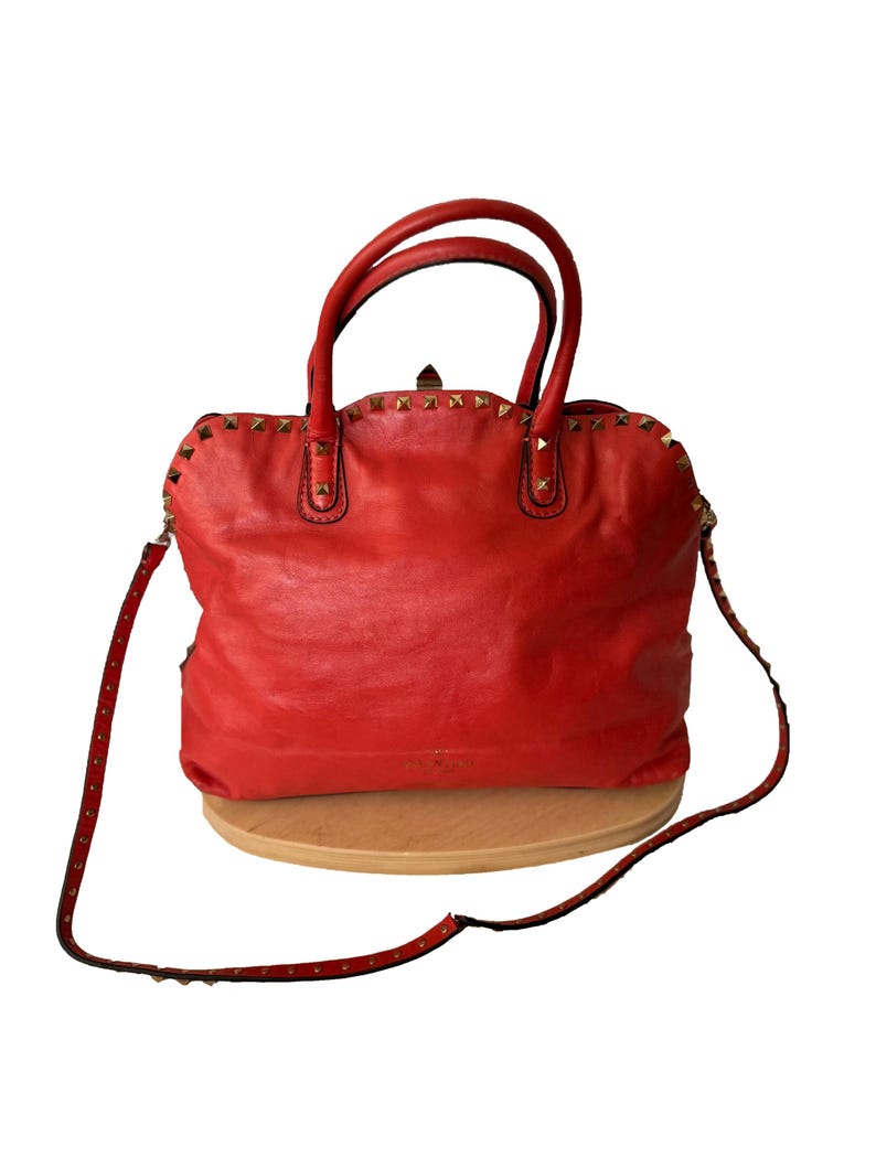 Peut inclure: Sac &agrave; main en cuir rouge vif orn&eacute; de clous dor&eacute;s sur le dessus et la bandouli&egrave;re. Il est dot&eacute; de deux poign&eacute;es sup&eacute;rieures et d'une bandouli&egrave;re amovible. Le sac est pos&eacute; sur une surface en bois.