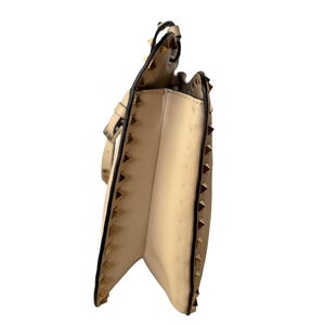 Pu&ograve; includere: Una borsa beige con borchie piramidali color oro lungo i bordi e un bordo nero. La borsa ha una chiusura con patta e una tracolla. L'interno &egrave; di colore beige chiaro.