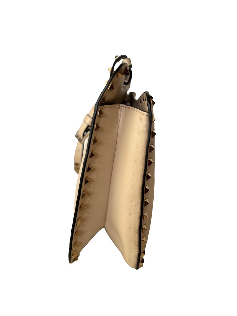 Pu&ograve; includere: Una borsa beige con borchie piramidali color oro lungo i bordi e un bordo nero. La borsa ha una chiusura con patta e una tracolla. L'interno &egrave; di colore beige chiaro.