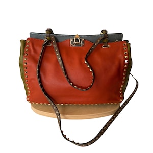 Puede incluir: Un bolso de mano de cuero con una combinaci&oacute;n de colores vibrantes. El bolso presenta un centro naranja quemado, laterales verde oliva y una parte superior gris. Tiene una correa con tachuelas y herrajes dorados. El bolso tiene un asa superior y una bandolera desmontable.