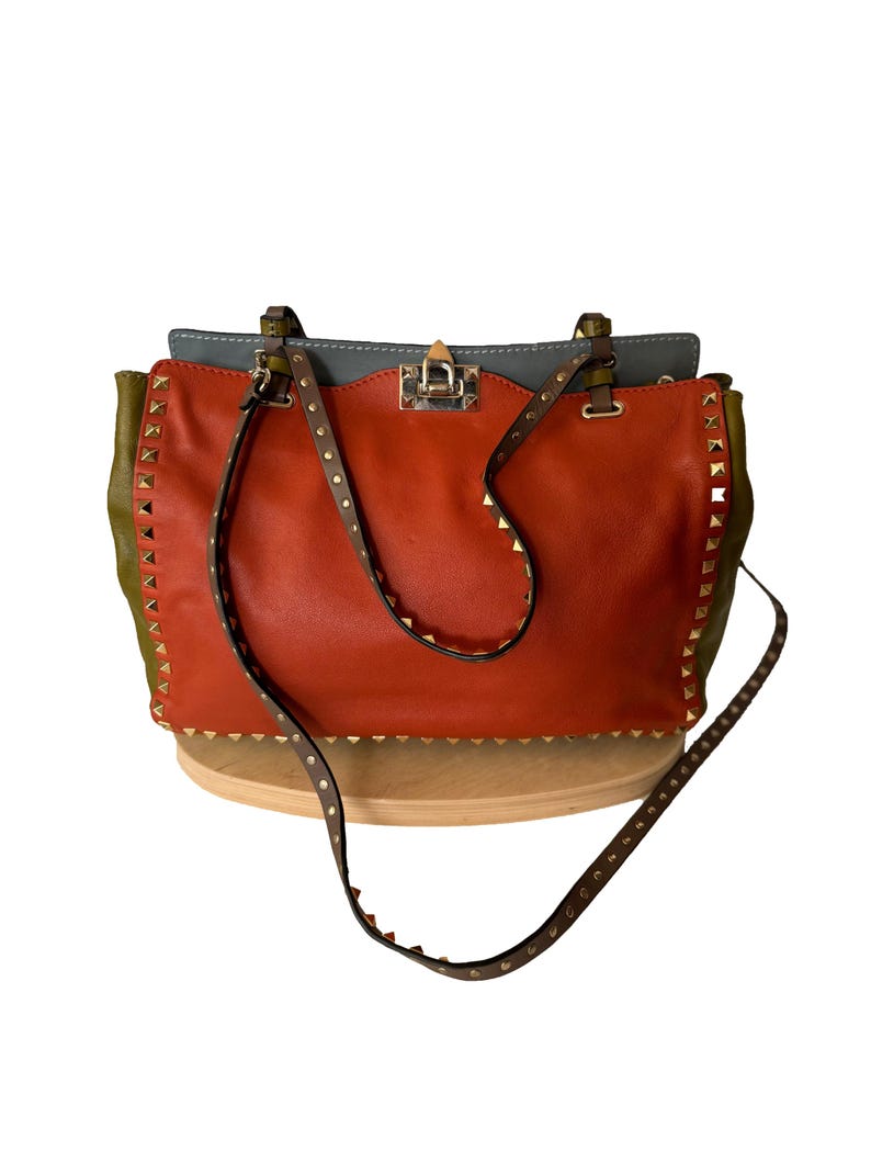 Puede incluir: Un bolso de mano de cuero con una combinaci&oacute;n de colores vibrantes. El bolso presenta un centro naranja quemado, laterales verde oliva y una parte superior gris. Tiene una correa con tachuelas y herrajes dorados. El bolso tiene un asa superior y una bandolera desmontable.