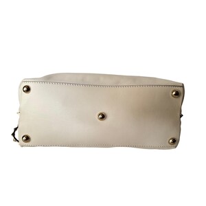 Puede incluir: Bolso de mano color crema con pies y tachuelas de metal dorado. Se ve la parte inferior del bolso, mostrando los detalles de las costuras y los herrajes. Un accesorio elegante para el uso diario.