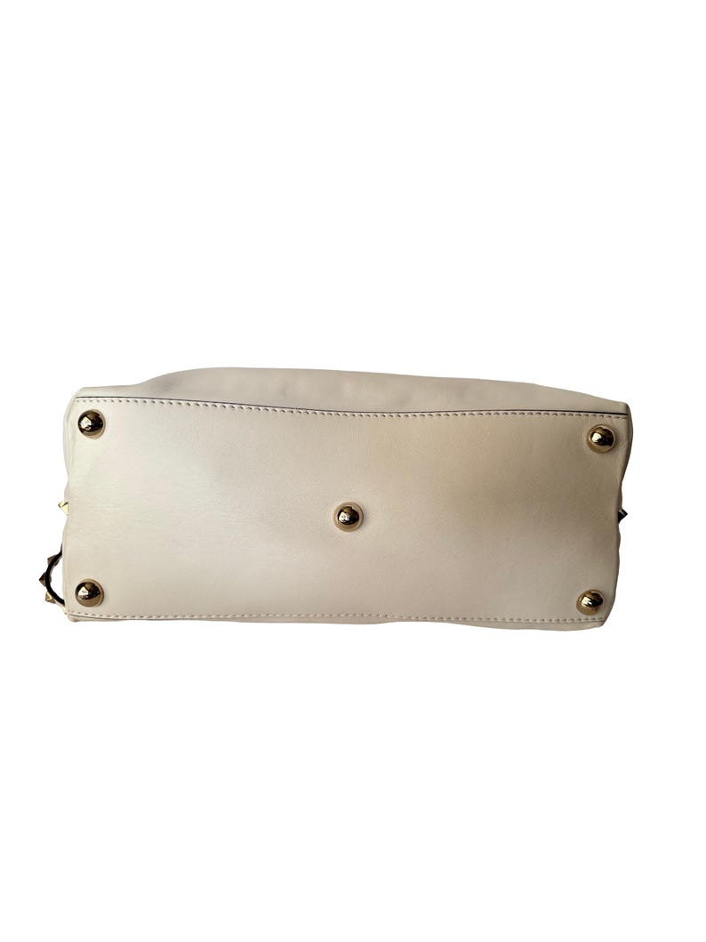 Puede incluir: Bolso de mano color crema con pies y tachuelas de metal dorado. Se ve la parte inferior del bolso, mostrando los detalles de las costuras y los herrajes. Un accesorio elegante para el uso diario.