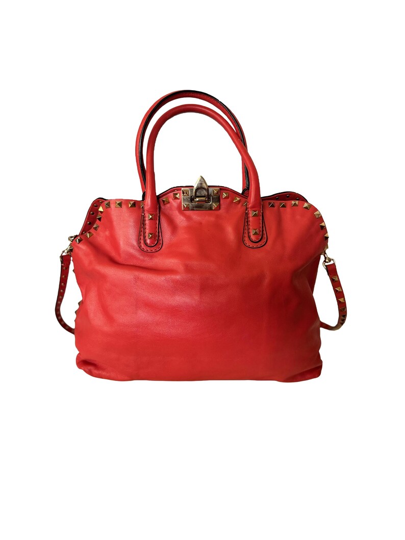 Peut inclure: Un sac &agrave; main en cuir rouge vif avec des clous dor&eacute;s. Le sac est dot&eacute; de deux poign&eacute;es sup&eacute;rieures et d'une bandouli&egrave;re amovible. La quincaillerie est dor&eacute;e et le sac a une forme classique et structur&eacute;e.