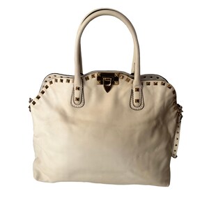 Puede incluir: Bolso de mano de cuero color crema con tachuelas doradas. El bolso tiene dos asas superiores y una forma rectangular con cierre de solapa. Las tachuelas est&aacute;n colocadas en la parte superior y los lados del bolso.