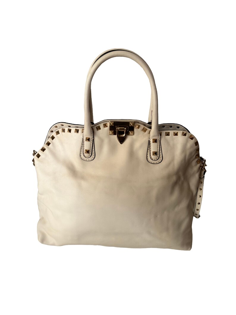 Puede incluir: Bolso de mano de cuero color crema con tachuelas doradas. El bolso tiene dos asas superiores y una forma rectangular con cierre de solapa. Las tachuelas est&aacute;n colocadas en la parte superior y los lados del bolso.