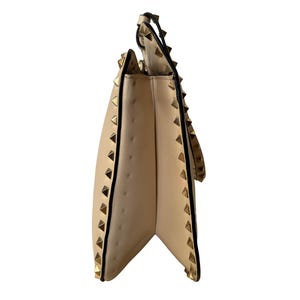 Pu&ograve; includere: Borsa a mano in pelle beige con borchie piramidali color oro lungo i bordi e il cinturino. La borsa ha una chiusura con patta e un bordo nero. Vista laterale della borsa.