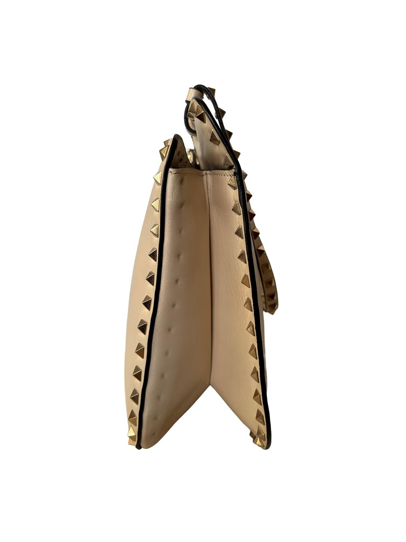 Pu&ograve; includere: Borsa a mano in pelle beige con borchie piramidali color oro lungo i bordi e il cinturino. La borsa ha una chiusura con patta e un bordo nero. Vista laterale della borsa.