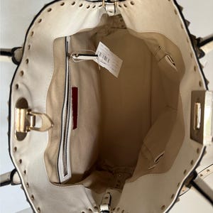 Puede incluir: Bolso de mano color crema con herrajes dorados y ribete con tachuelas. El interior es beige con un bolsillo con cremallera y un acento rojo. El bolso tiene un asa superior y una bandolera.