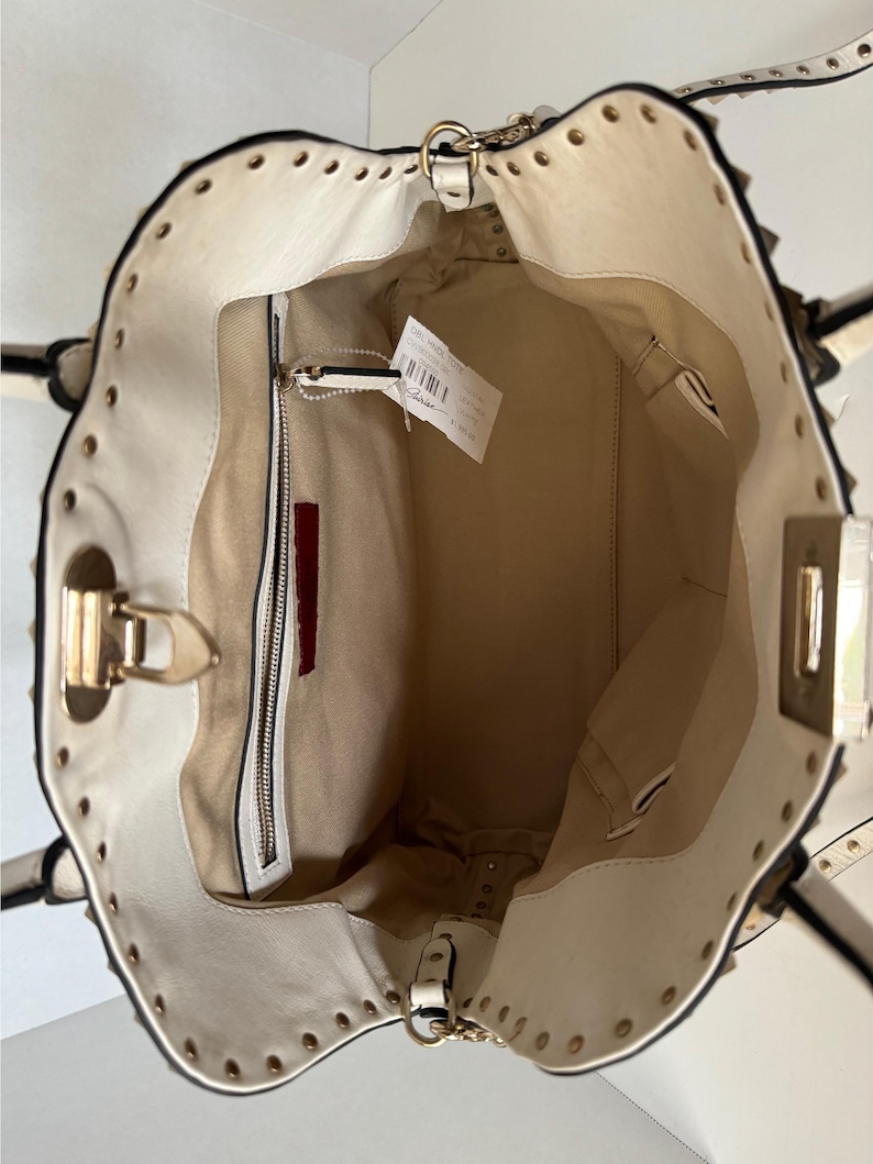 Puede incluir: Bolso de mano color crema con herrajes dorados y ribete con tachuelas. El interior es beige con un bolsillo con cremallera y un acento rojo. El bolso tiene un asa superior y una bandolera.