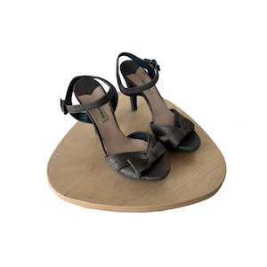 May include: A pair of dark gray high-heeled sandals with ankle straps and bow details. The sandals are displayed on a light brown, wooden, oval-shaped surface. The heels are approximately 3 inches tall.