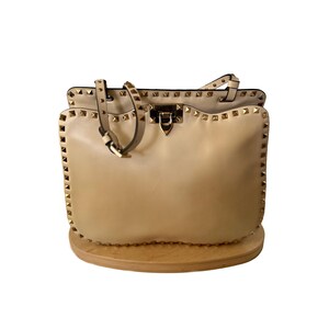 Pu&ograve; includere: Una borsa in pelle beige con borchie color oro lungo i bordi e i cinturini. La borsa ha un manico superiore e una chiusura con fibbia. La borsa &egrave; appoggiata su un espositore in legno.