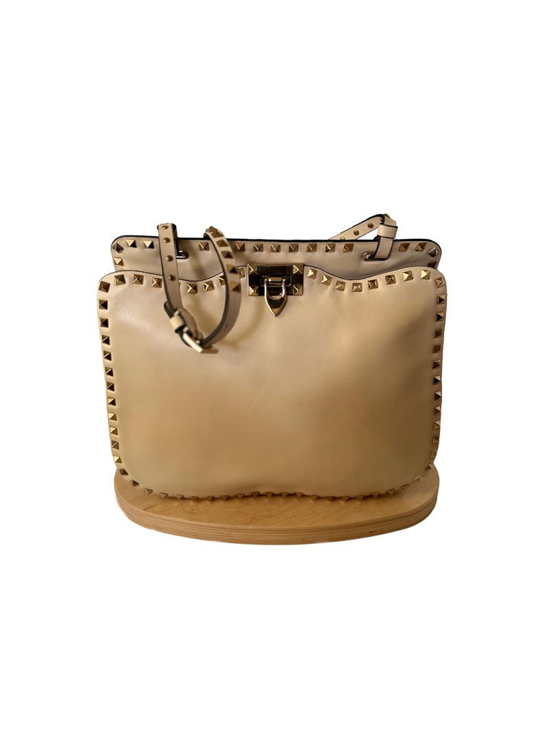 Pu&ograve; includere: Una borsa in pelle beige con borchie color oro lungo i bordi e i cinturini. La borsa ha un manico superiore e una chiusura con fibbia. La borsa &egrave; appoggiata su un espositore in legno.