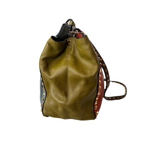 Puede incluir: Un bolso tipo cubo de cuero multicolor con cierre de cord&oacute;n. El bolso presenta paneles verde oliva, rojos y azules, acentuados con tachuelas doradas. El bolso tiene una correa de cuero marr&oacute;n.