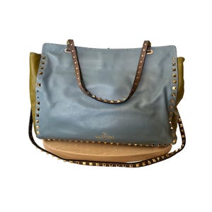 Puede incluir: Un bolso tote de cuero azul con correas marrones y tachuelas doradas. El bolso tiene forma rectangular con una parte delantera azul claro y laterales verde oliva. La marca "Valentino" es visible.