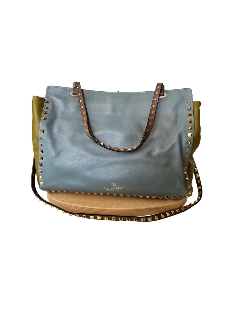 Puede incluir: Un bolso tote de cuero azul con correas marrones y tachuelas doradas. El bolso tiene forma rectangular con una parte delantera azul claro y laterales verde oliva. La marca "Valentino" es visible.