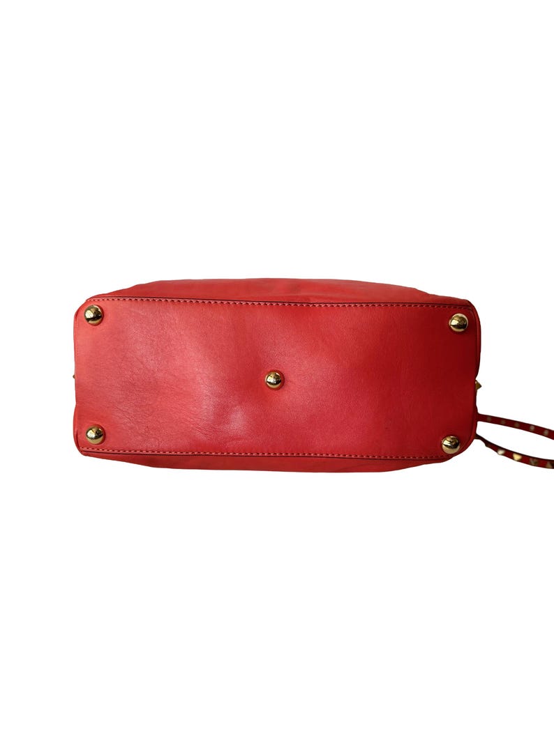 Peut inclure: Sac &agrave; main en cuir rouge avec des clous dor&eacute;s sur le fond. Le sac est de forme rectangulaire avec des coins arrondis. La base du sac est visible, avec des coutures et des &eacute;l&eacute;ments m&eacute;talliques.