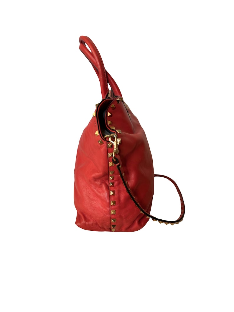 Peut inclure: Un sac &agrave; main en cuir rouge avec des clous dor&eacute;s. Le sac a une poign&eacute;e sup&eacute;rieure et une bandouli&egrave;re amovible. Le sac est fait d'un cuir souple et textur&eacute;.