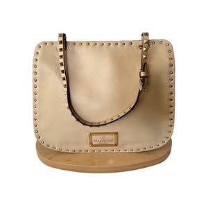 Pu&ograve; includere: Borsa a mano in pelle beige con borchie color oro sui bordi e sui cinturini. La borsa ha una forma rettangolare con un manico superiore e una piccola targhetta con logo rettangolare.
