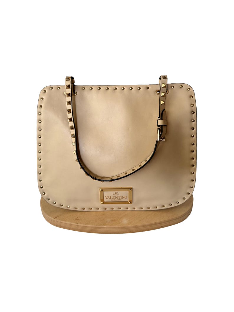 Pu&ograve; includere: Borsa a mano in pelle beige con borchie color oro sui bordi e sui cinturini. La borsa ha una forma rettangolare con un manico superiore e una piccola targhetta con logo rettangolare.