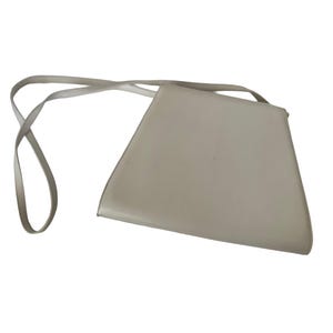 Puede incluir: Un bolso cruzado de color gris claro, con forma de trapecio y una correa larga y fina. El bolso tiene un dise&ntilde;o minimalista y parece estar hecho de un material liso. La correa est&aacute; sujeta a las esquinas superiores del bolso.