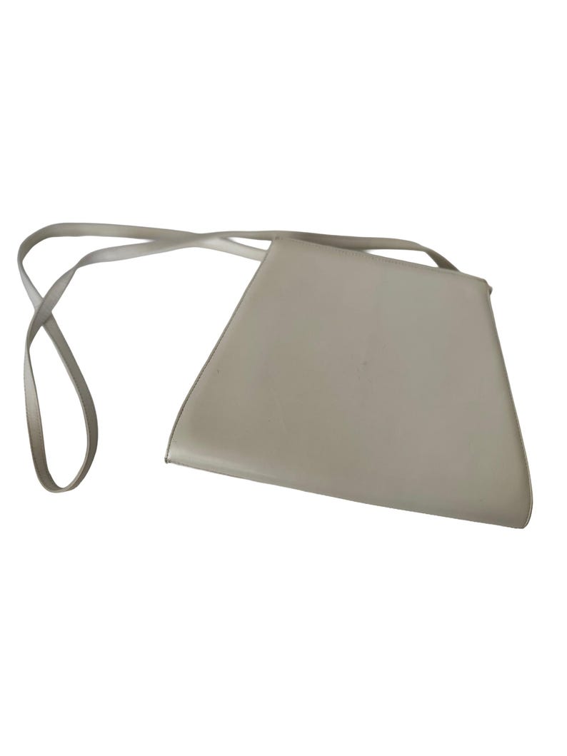 Puede incluir: Un bolso cruzado de color gris claro, con forma de trapecio y una correa larga y fina. El bolso tiene un dise&ntilde;o minimalista y parece estar hecho de un material liso. La correa est&aacute; sujeta a las esquinas superiores del bolso.