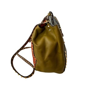 Puede incluir: Mochila de cuero verde oliva con correas marrones y tachuelas doradas. La mochila tiene un cierre de solapa y una secci&oacute;n estampada con detalles en rojo y gris. Las correas son ajustables.