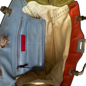 Puede incluir: Vista abierta de un bolso de mano de cuero multicolor. El bolso presenta secciones de cuero azul, verde oliva y naranja. El interior es beige con un bolsillo azul y una etiqueta roja. Se ven detalles de tachuelas y herrajes met&aacute;licos.