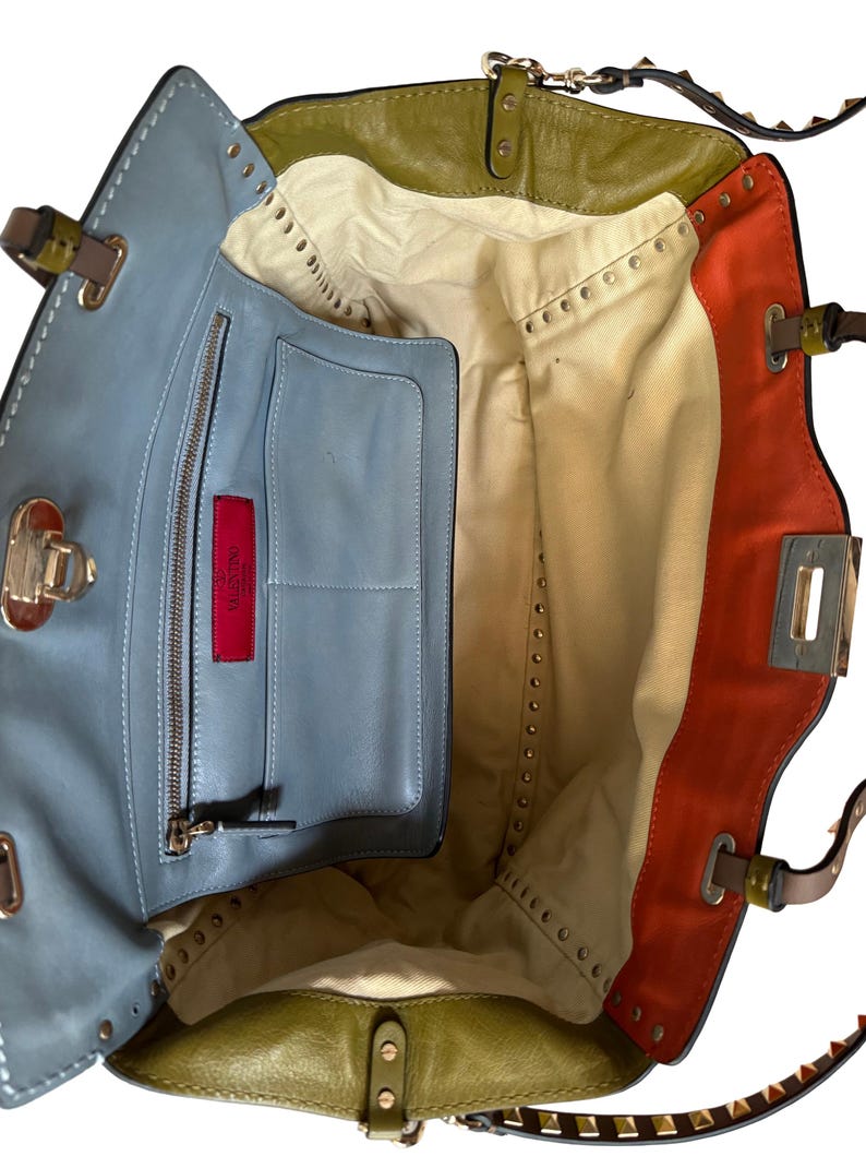 Puede incluir: Vista abierta de un bolso de mano de cuero multicolor. El bolso presenta secciones de cuero azul, verde oliva y naranja. El interior es beige con un bolsillo azul y una etiqueta roja. Se ven detalles de tachuelas y herrajes met&aacute;licos.