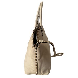 Puede incluir: Bolso de mano color crema con tachuelas piramidales doradas en los laterales. El bolso tiene dos asas y una correa para el hombro desmontable. El bolso est&aacute; hecho de un material suave y texturizado.