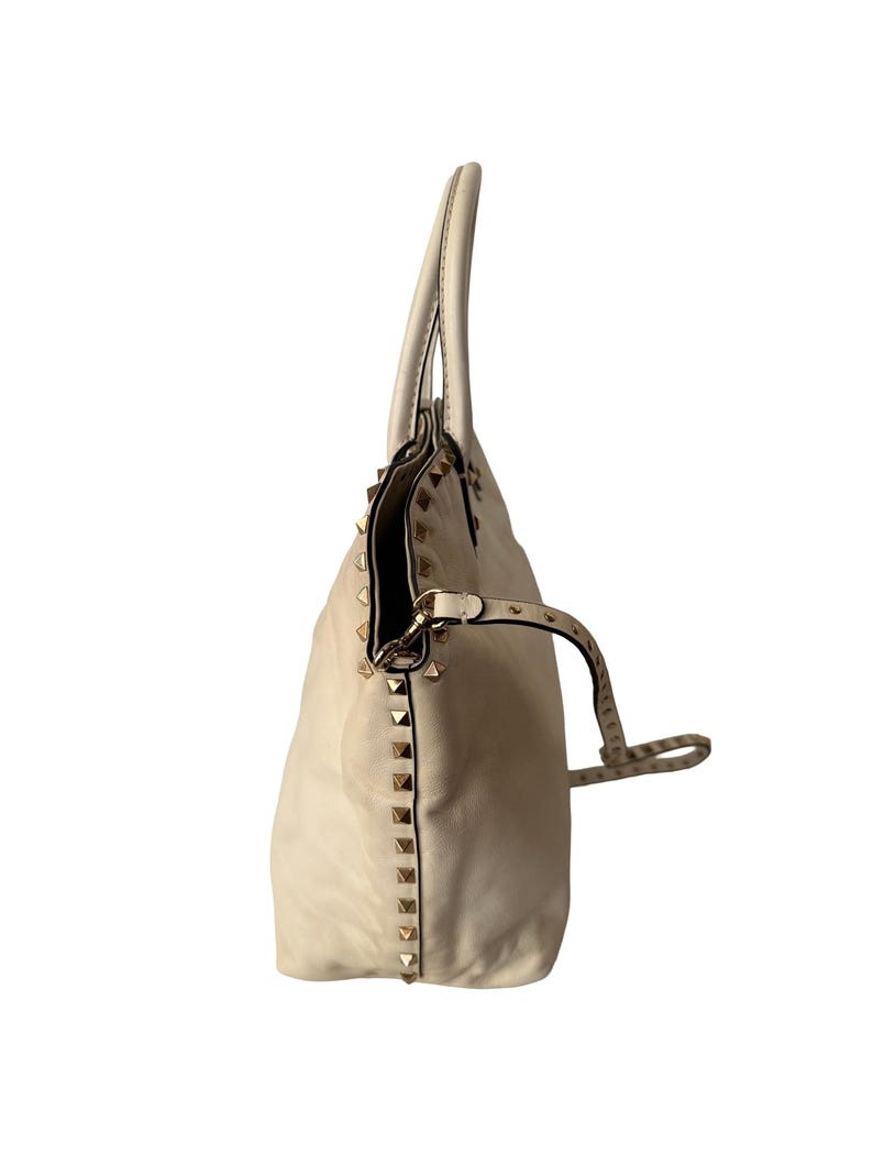 Puede incluir: Bolso de mano color crema con tachuelas piramidales doradas en los laterales. El bolso tiene dos asas y una correa para el hombro desmontable. El bolso est&aacute; hecho de un material suave y texturizado.