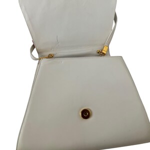 Puede incluir: Un bolso blanco en forma de trapecio con asa superior y bandolera. El bolso tiene un cierre y herrajes dorados. El interior es visible y el bolso parece estar hecho de cuero o material similar. El bolso est&aacute; abierto, revelando el interior.