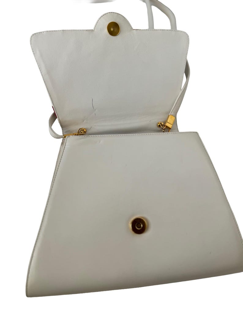 Puede incluir: Un bolso blanco en forma de trapecio con asa superior y bandolera. El bolso tiene un cierre y herrajes dorados. El interior es visible y el bolso parece estar hecho de cuero o material similar. El bolso est&aacute; abierto, revelando el interior.