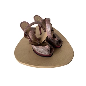 May include: A pair of pink platform high heels with a snakeskin pattern. The open-toe shoes have thin ankle straps and are displayed on a light brown, triangular wooden surface.