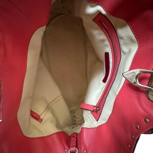 Peut inclure: Vue ouverte d'un sac fourre-tout en cuir rouge avec des rivets dor&eacute;s. L'int&eacute;rieur pr&eacute;sente une doublure beige et une poche zipp&eacute;e. Le sac a un fermoir argent&eacute; et un petit accent en cuir rouge.