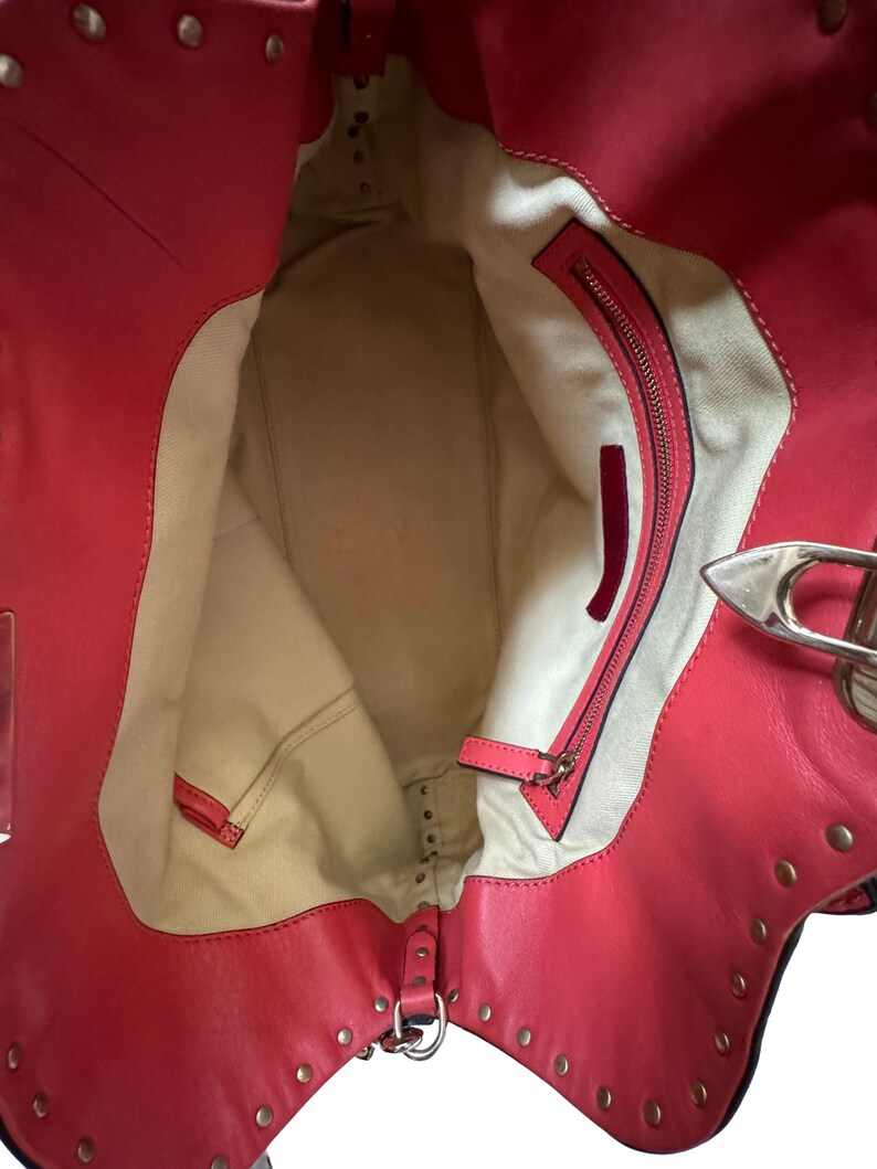 Peut inclure: Vue ouverte d'un sac fourre-tout en cuir rouge avec des rivets dor&eacute;s. L'int&eacute;rieur pr&eacute;sente une doublure beige et une poche zipp&eacute;e. Le sac a un fermoir argent&eacute; et un petit accent en cuir rouge.
