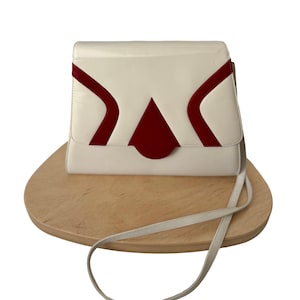 Puede incluir: Un bolso de mano color crema con un dise&ntilde;o geom&eacute;trico en rojo. El bolso tiene un cierre de solapa y una correa larga y fina. El bolso est&aacute; sobre una superficie de madera.