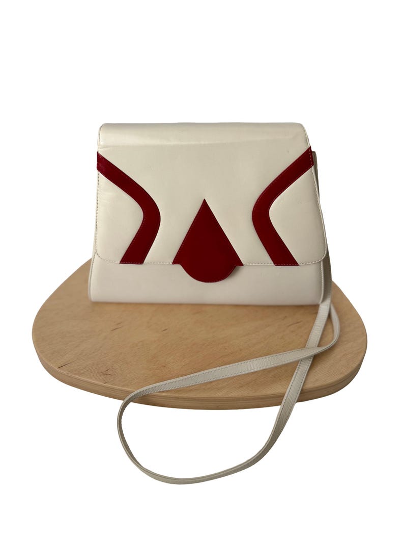 Puede incluir: Un bolso de mano color crema con un dise&ntilde;o geom&eacute;trico en rojo. El bolso tiene un cierre de solapa y una correa larga y fina. El bolso est&aacute; sobre una superficie de madera.