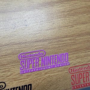 Op de afbeelding: Een paarse sticker met de tekst Nintendo Super Nintendo Entertainment System, een rode sticker met de tekst Nintendo Super Entertainment en een zwarte sticker met de tekst Nintendo Entertainment System op een houten oppervlak.