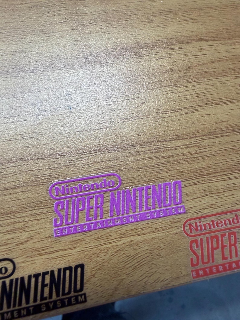Op de afbeelding: Een paarse sticker met de tekst Nintendo Super Nintendo Entertainment System, een rode sticker met de tekst Nintendo Super Entertainment en een zwarte sticker met de tekst Nintendo Entertainment System op een houten oppervlak.