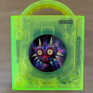 May include: A bright green Nintendo GameCube carry case. The case has a circular graphic of a mask with a red face, white spikes, and purple and blue accents. The Nintendo logo is visible on the top.