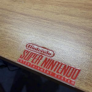 Op de afbeelding: Een lichtbruin houtnerfoppervlak met een glanzende, verhoogde rood-witte sticker met de tekst Nintendo Super Nintendo Entertainment System in een vet, gestileerd lettertype op het platte, gestructureerde materiaal.