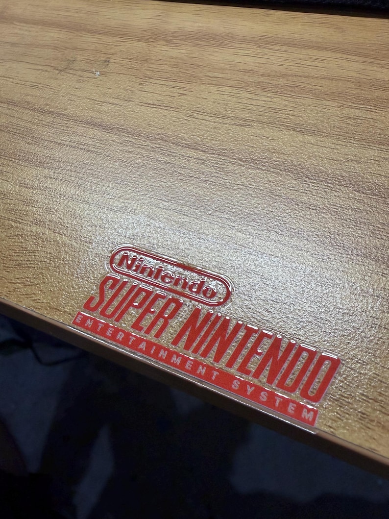 Op de afbeelding: Een lichtbruin houtnerfoppervlak met een glanzende, verhoogde rood-witte sticker met de tekst Nintendo Super Nintendo Entertainment System in een vet, gestileerd lettertype op het platte, gestructureerde materiaal.