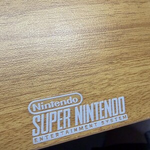 Op de afbeelding: Een close-up van een houten oppervlak met het witte Nintendo Super Nintendo Entertainment System logo. De houtnerf is zichtbaar en het oppervlak heeft een lichtbruine kleur.