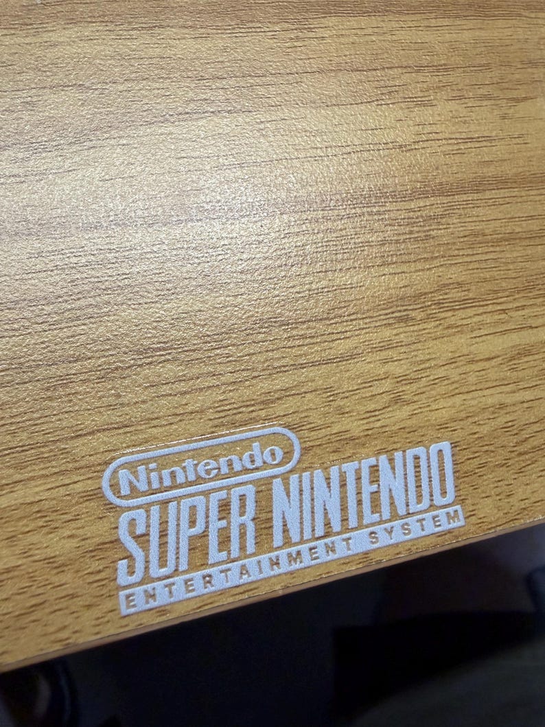 Op de afbeelding: Een close-up van een houten oppervlak met het witte Nintendo Super Nintendo Entertainment System logo. De houtnerf is zichtbaar en het oppervlak heeft een lichtbruine kleur.